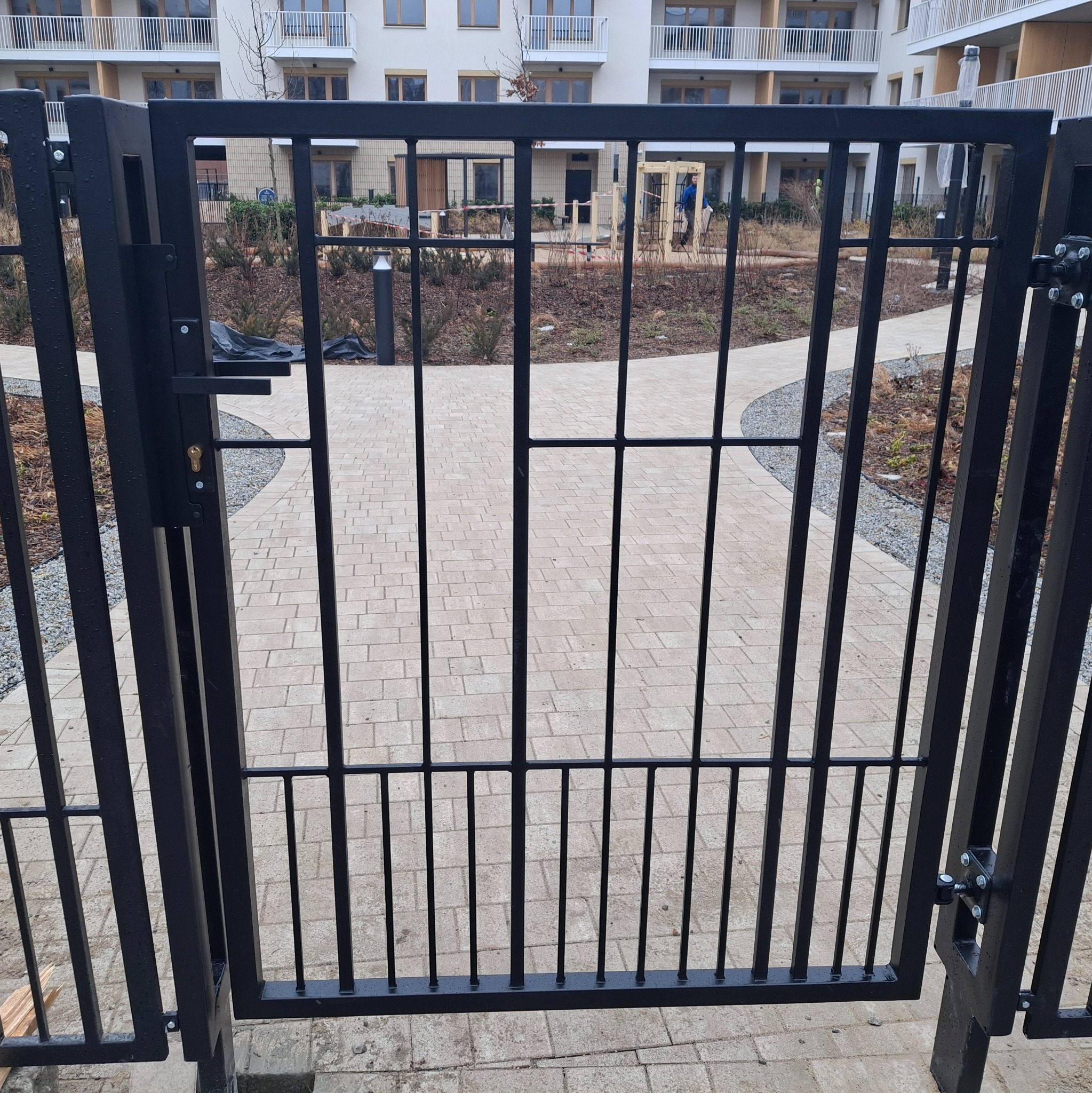 Otwarta, czarna, metalowa furtka z zamkiem, prowadząca do ogrodu z ścieżką z kostki brukowej. W tle budynek mieszkalny z balkonami.
