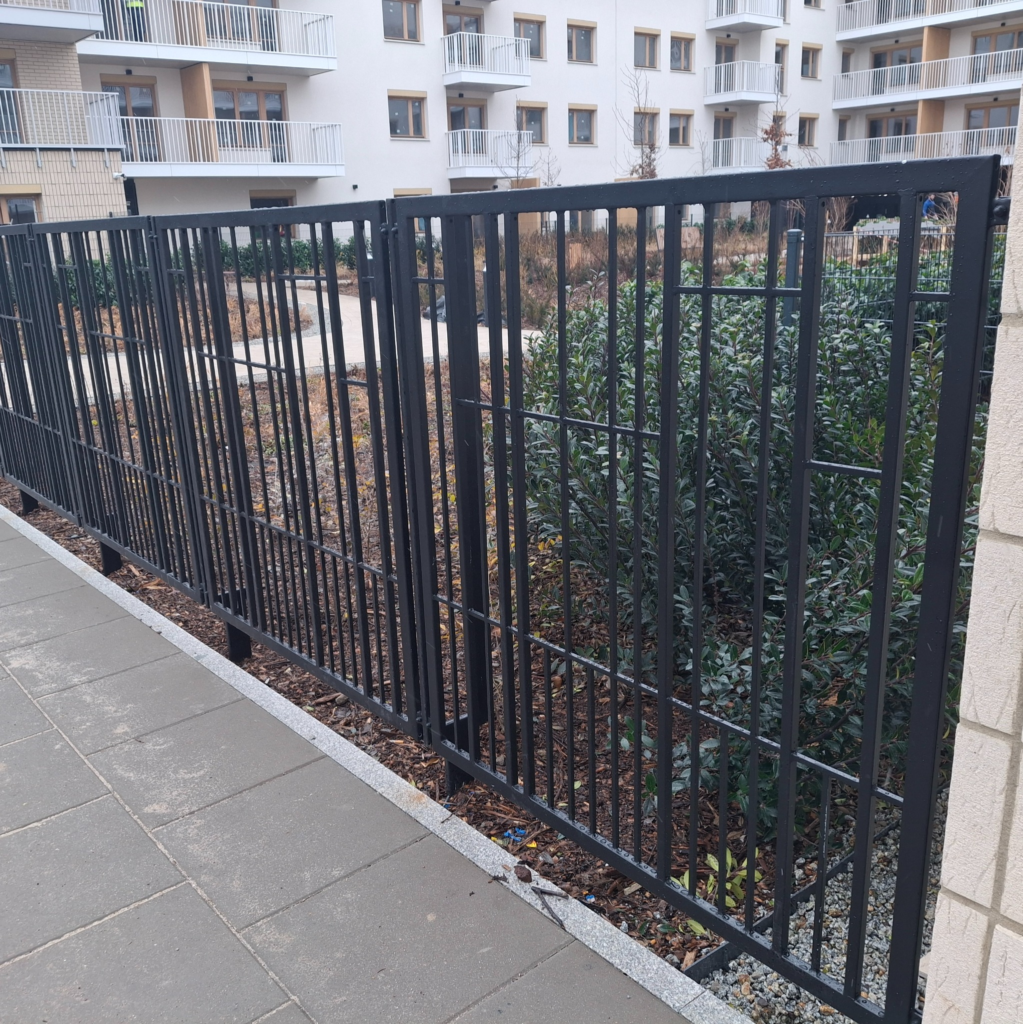 Czarne, metalowe ogrodzenie panelowe na tle nowoczesnego budynku mieszkalnego i zieleni. Widok z poziomu chodnika.