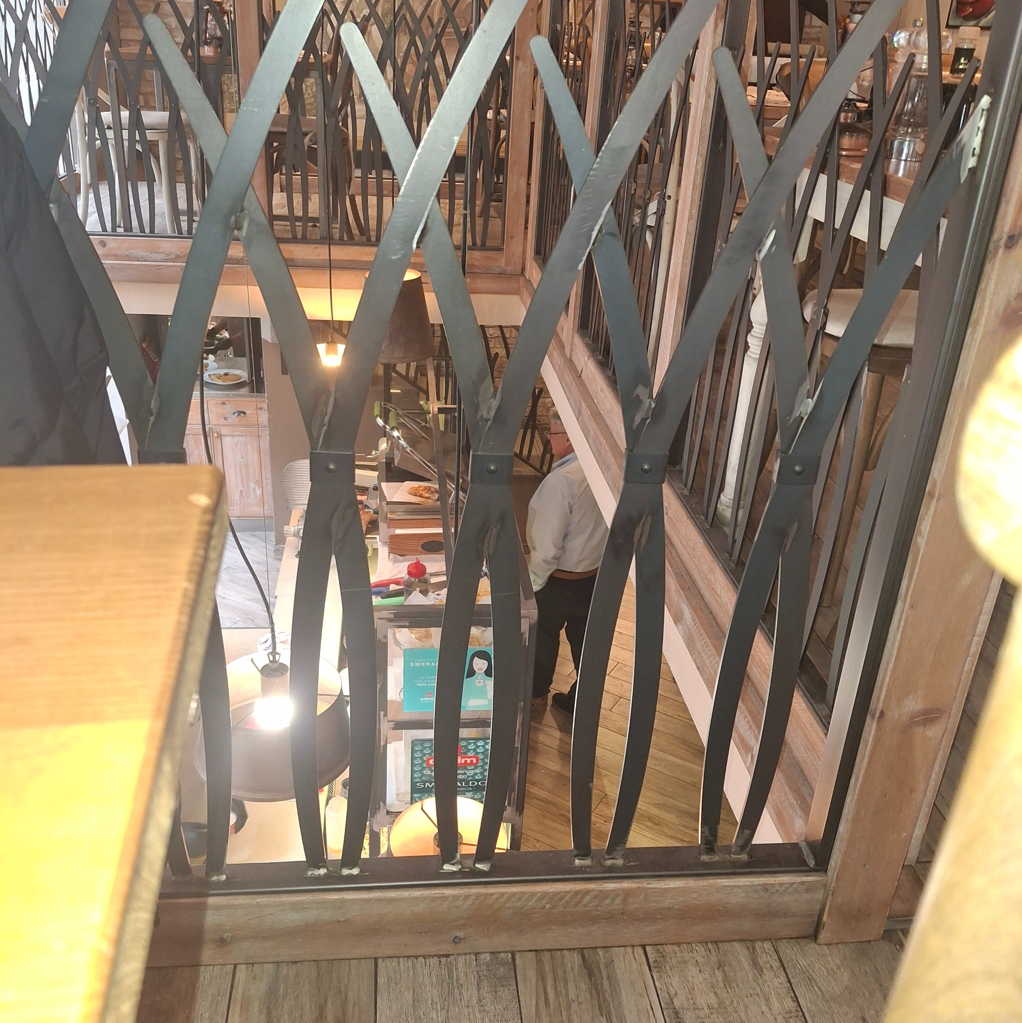 Metalowa balustrada o dekoracyjnym wzorze, widok z góry na wnętrze restauracji. Elementy spawane, drewniane wykończenie. Nietypowy kształt prętów.