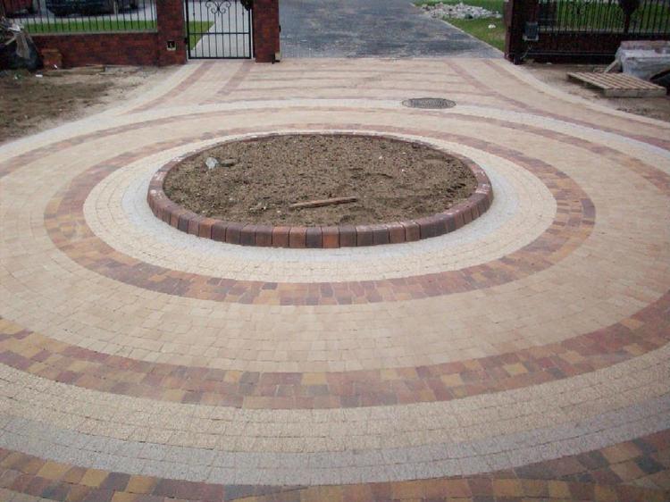 Okrągły wzór z kostki brukowej w odcieniach beżu i brązu tworzy podjazd z centralnym klombem. Brama w tle.