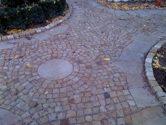 Ułożona z kostki granitowej ścieżka z okrągłym włazem w centrum i obrzeżami z jasnych kostek. Fragmenty liści na powierzchni. Krzewy w tle.