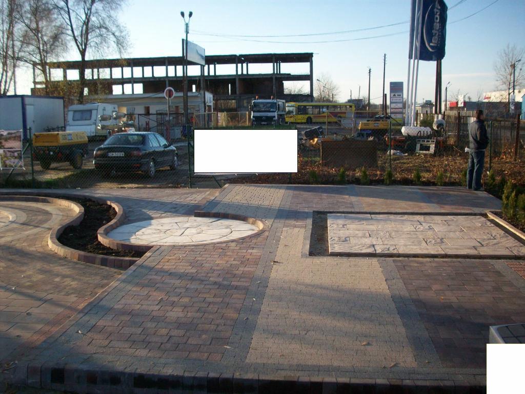 Wzorcowe ułożenie kostki brukowej w ogrodzie z elementami dekoracyjnymi: okrąg, kwadrat i falista rabata. W tle budynek w trakcie budowy i zaparkowane pojazdy.