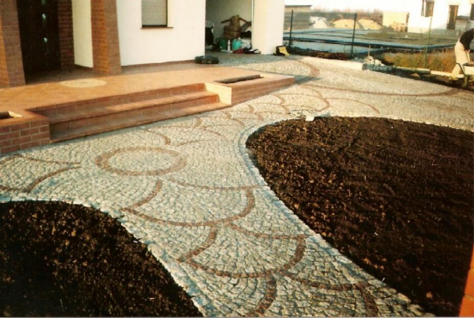 Granitowa ścieżka z dekoracyjnym wzorem ułożona przed domem, kontrastująca z ciemną ziemią. Schody wejściowe z cegły. Widok na posesję w tle.