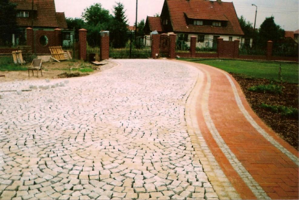 Granitowa kostka ułożona w łukowaty wzór, obok ścieżka z czerwonej kostki, na tle domu z czerwonej cegły i zieleni. Widok z perspektywy chodnika.