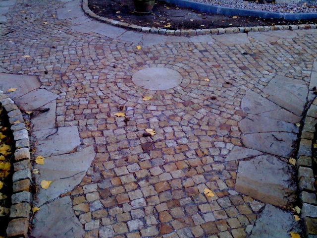 Ułożona kostka granitowa w kształcie koła, przechodząca w ścieżkę z płyt kamiennych, z jesiennymi liśćmi. Widok z góry na wykończony fragment ogrodu.