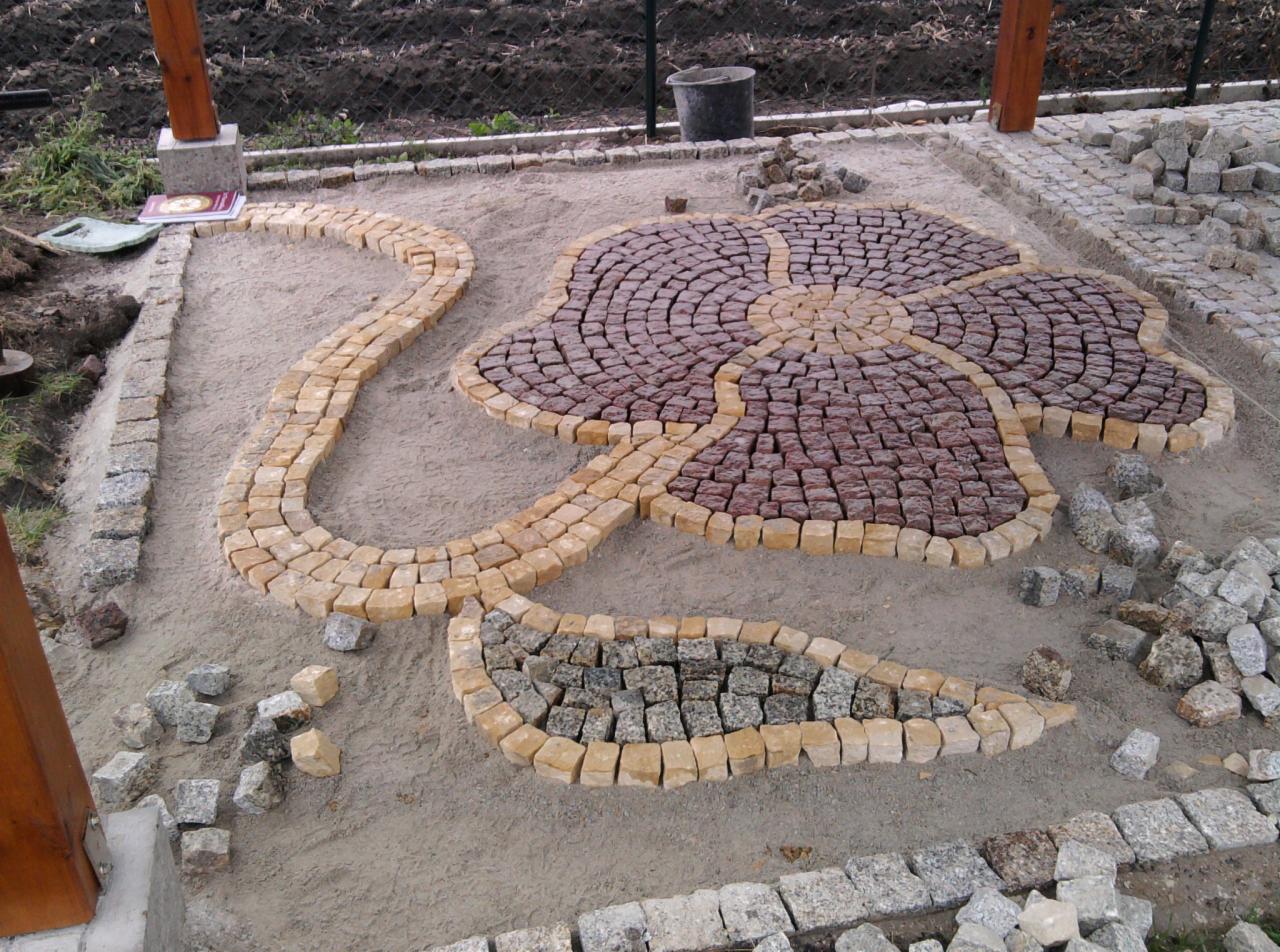 Ozdobny wzór kwiatowy z kostki granitowej w odcieniach brązu i beżu, w trakcie układania. Widoczne nierówności podłoża i porozrzucane kostki, co sugeruje etap prac.