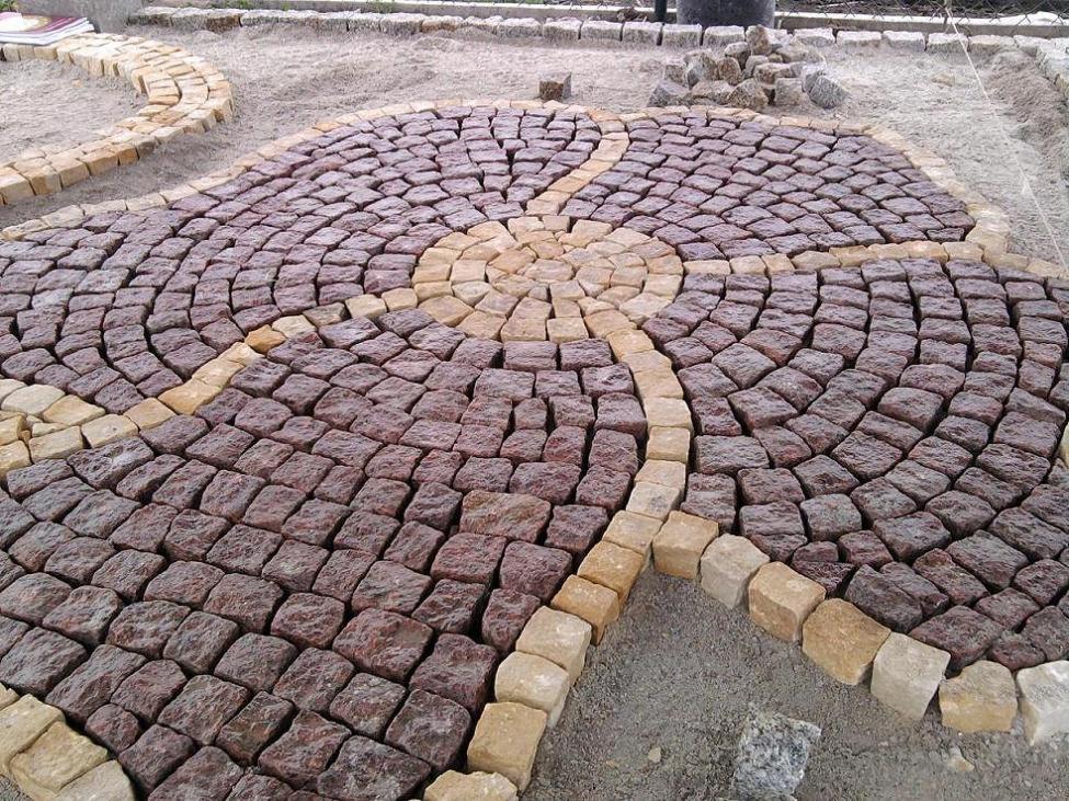 Okrągła powierzchnia z brązowo-żółtej kostki granitowej ułożonej w dekoracyjny wzór słoneczny, otoczona piaskiem i częściowo ułożonym chodnikiem.