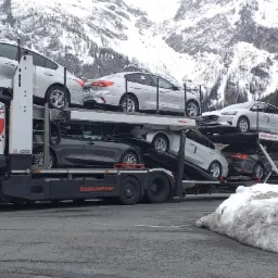 Ciężarówka z lawetą marki Kässbohrer przewożąca kilka samochodów osobowych na tle gór pokrytych śniegiem.
