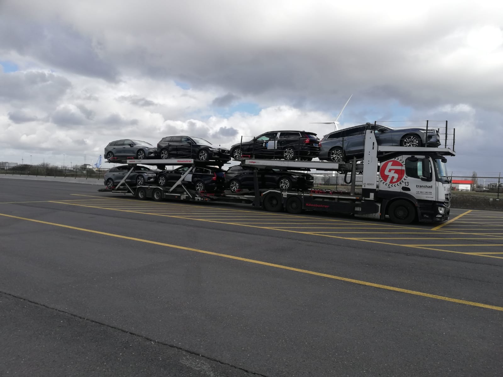 Laweta z ośmioma samochodami Volvo na parkingu lotniskowym z wiatrakiem w tle, pochmurne niebo.
