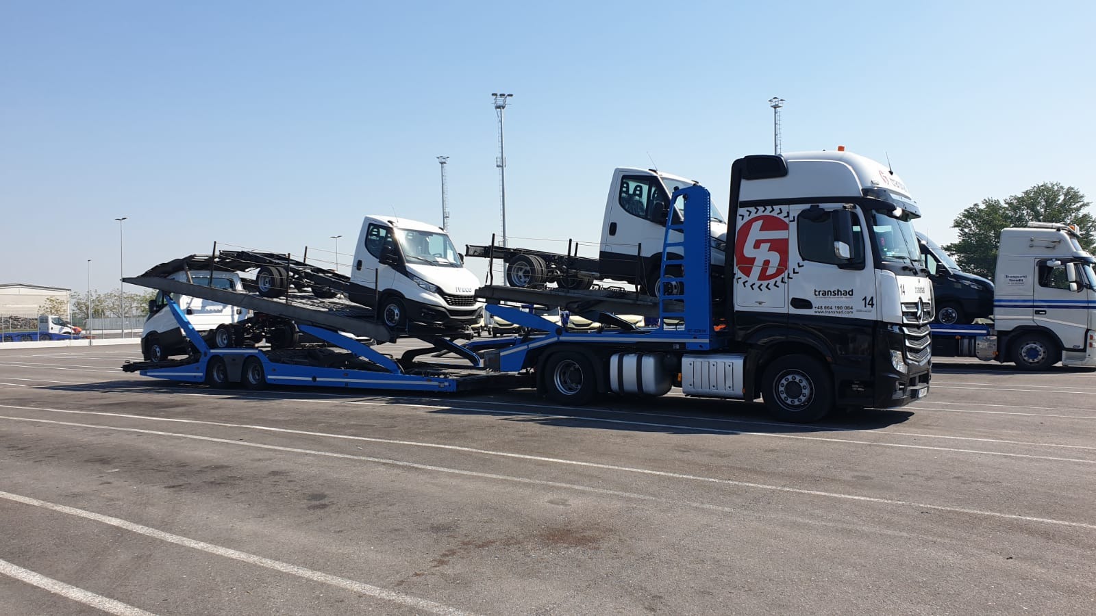 Ciężarówka z lawetą przewożąca dwa białe samochody dostawcze marki Iveco na parkingu, z widocznym logo firmy transportowej 'transhad' na kabinie.