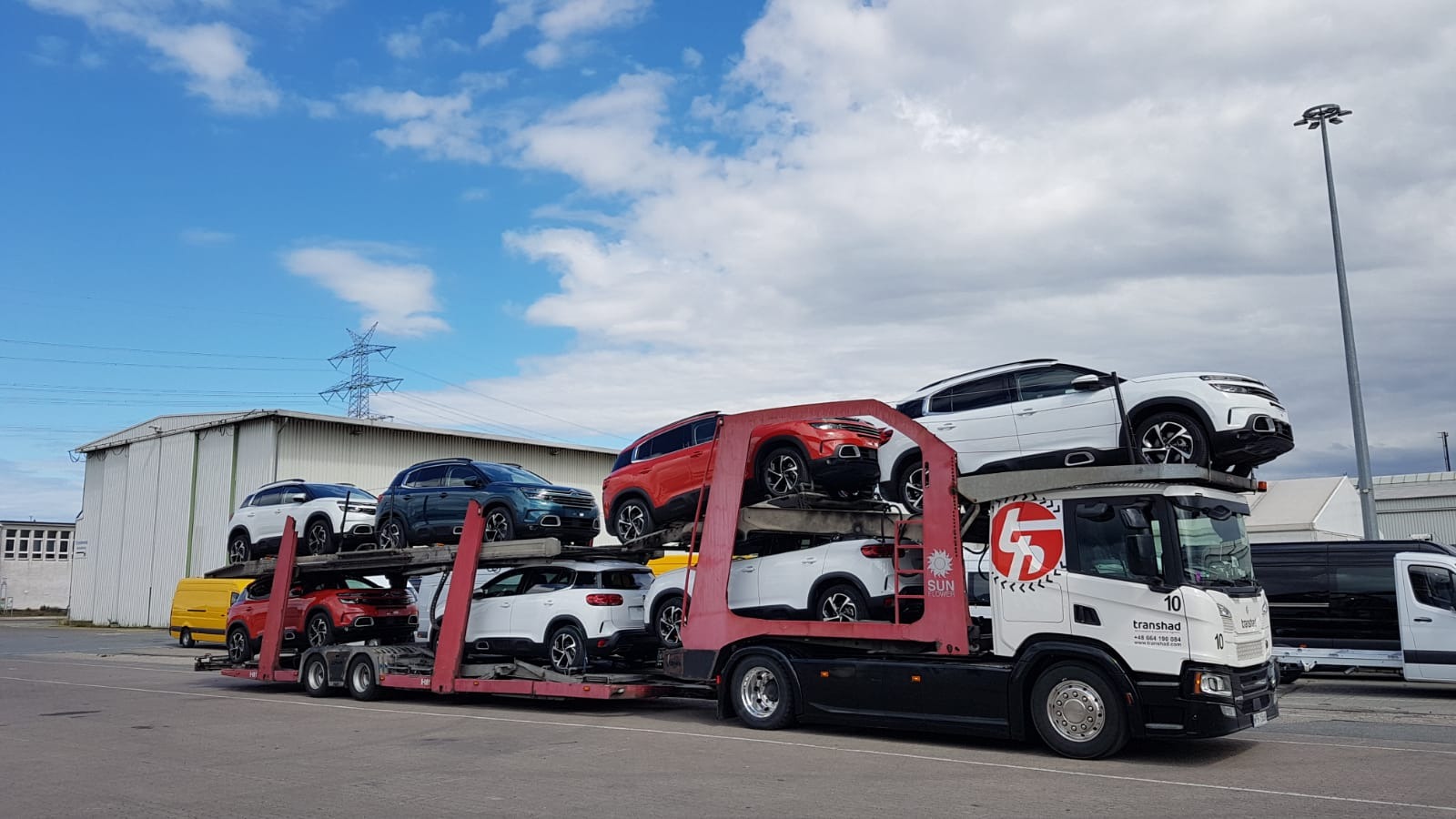 Ciężarówka laweta przewożąca kilka samochodów osobowych różnych kolorów na tle błękitnego nieba z chmurami i zabudowań przemysłowych.