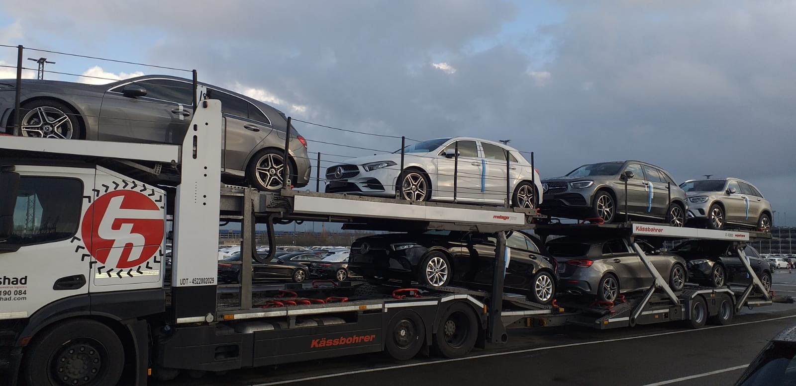 Laweta marki Kässbohrer z logo firmy transportowej SHAD przewozi nowe samochody marki Mercedes-Benz z niebieskimi paskami zabezpieczającymi na tle parkingu z dużą ilością aut.
