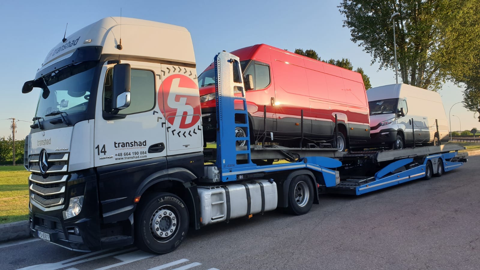 Ciężarówka laweta marki Mercedes z logo firmy Transhad przewozi dwa busy: czerwony i biały. Widok z boku, w tle drzewa i błękitne niebo.