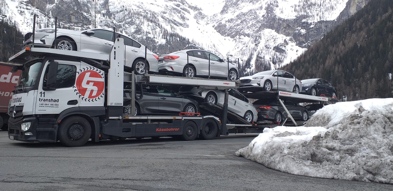 Ciężarówka z lawetą marki Kässbohrer przewożąca kilka samochodów osobowych na tle gór pokrytych śniegiem.