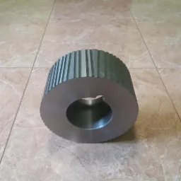 Szara, metalowa rolka z rowkami i otworem, leżąca na kafelkach. Precyzyjnie wykonany element, prawdopodobnie po obróbce skrawaniem.