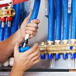 Instalator podłącza niebieski przewód do mosiężnego rozdzielacza z niebieskimi nakrętkami, widoczne inne przewody oraz elementy instalacji.