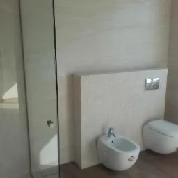 Wykończona łazienka z kabiną prysznicową typu walk-in, bidetem i toaletą. Jasne płytki na ścianach i podłoga imitująca drewno. Minimalistyczny design z chromowanymi elementami.