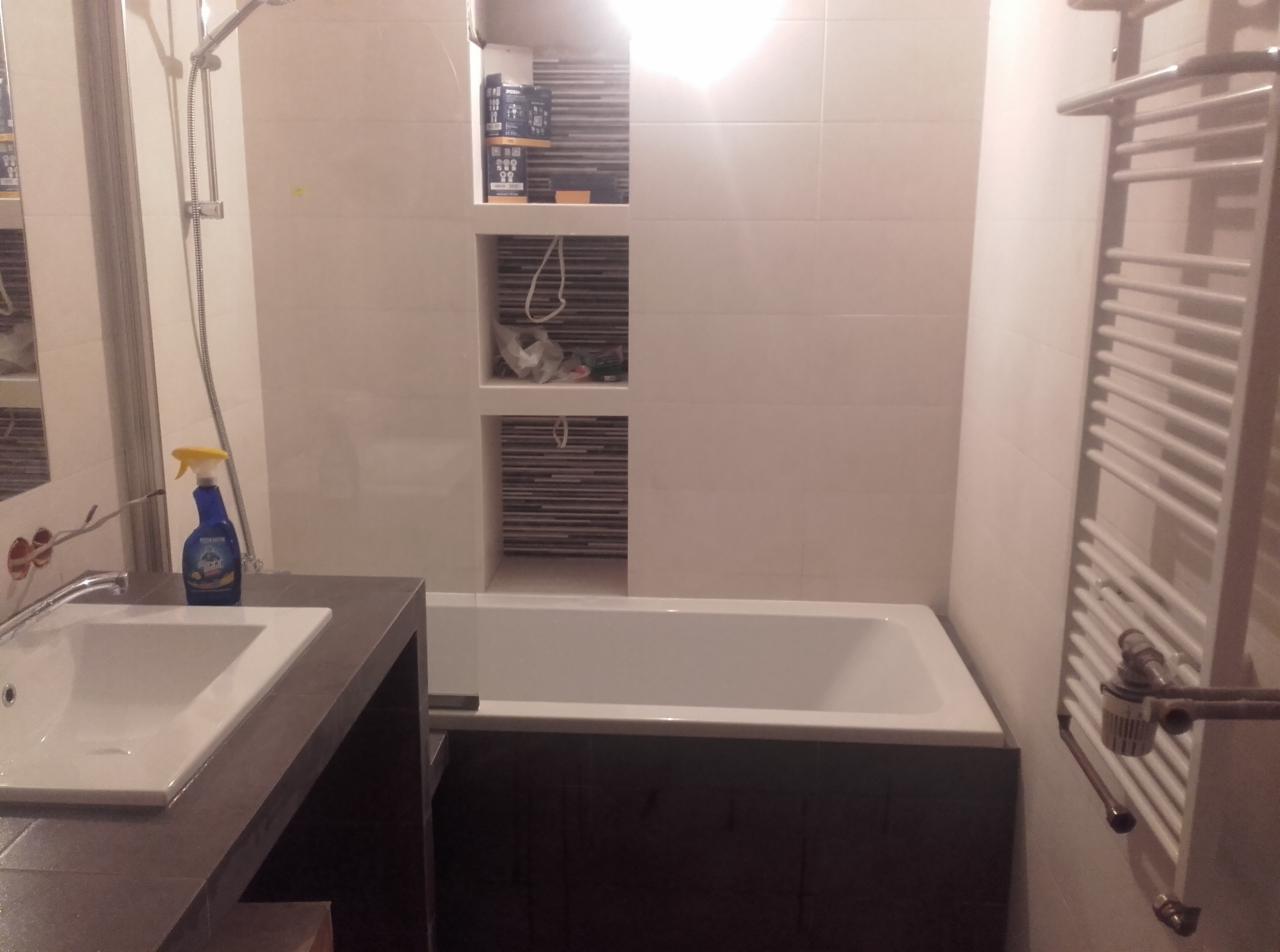 Wykończona łazienka z białą wanną obudowaną ciemnobrązowymi panelami, białą umywalką z szarą szafką, grzejnikiem drabinkowym i wnękami w ścianie wyłożonymi mozaiką.