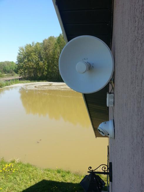 Biała antena satelitarna przymocowana do ściany budynku, z widokiem na zarośnięty staw i drzewa w tle.