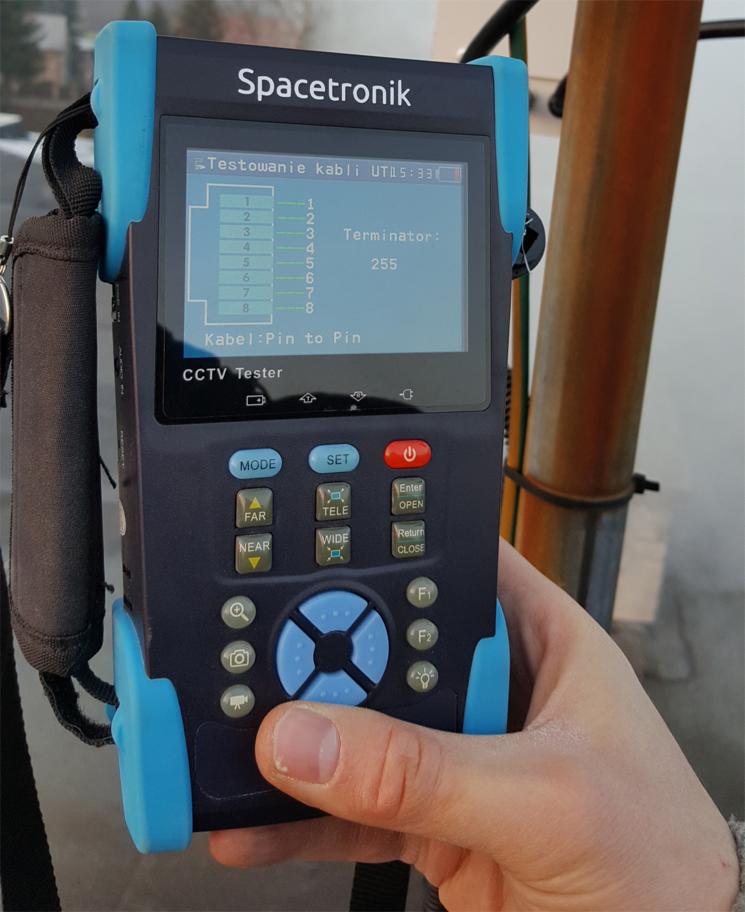 Tester kabli Spacetronik w dłoni, wyświetlający wynik testu okablowania, prawdopodobnie CCTV, na tle konstrukcji stalowej.