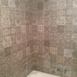 Ściana w łazience wyłożona szarymi, wzorzystymi płytkami ceramicznymi nad wanną, widoczne wystające rurki instalacji wodnej i fragmenty kabli elektrycznych pod sufitem.