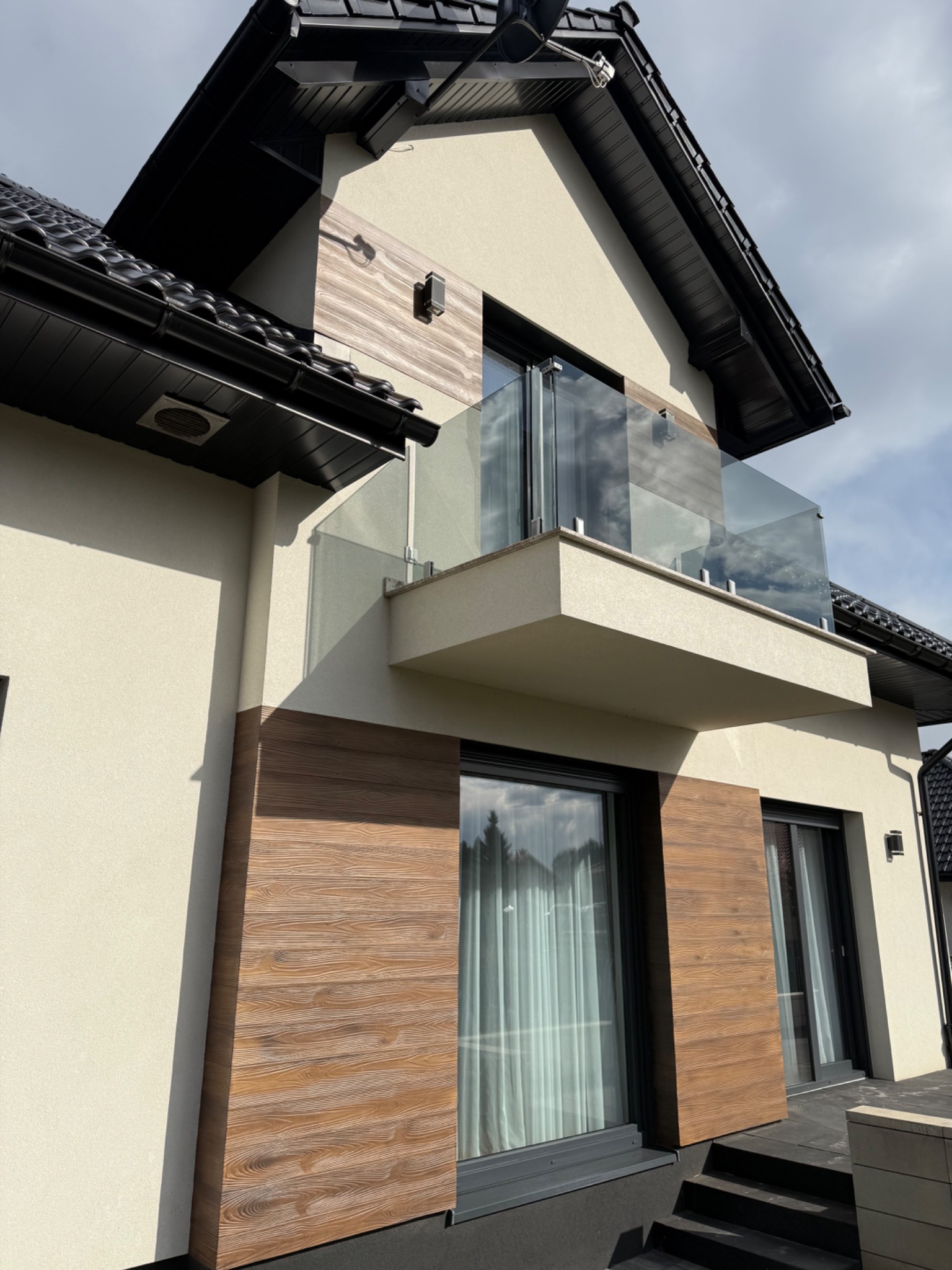 Nowoczesny dom z balkonem i drewnianą elewacją. Jasna fasada, czarne okna i dach. Balustrada balkonowa ze szkła. Ujęcie z dołu, podkreślające architekturę budynku.