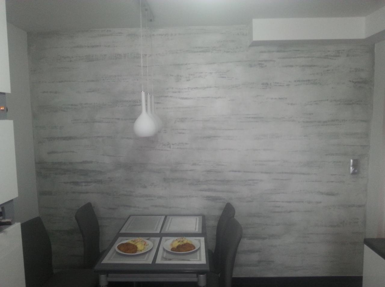 Ściana w jadalni z dekoracyjnym tynkiem o fakturze przypominającej poziome smugi, oświetlona wiszącą lampą z trzema białymi kloszami, stół z dwoma nakrytymi miejscami i posiłkami.