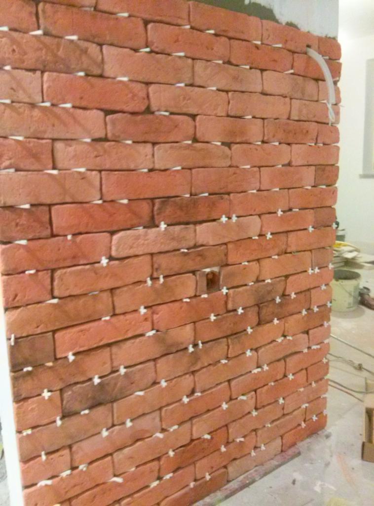 Ściana wyłożona cegłami klinkierowymi w trakcie montażu, z krzyżykami dystansowymi w spoinach, widoczna rurka instalacyjna przy górnej krawędzi.