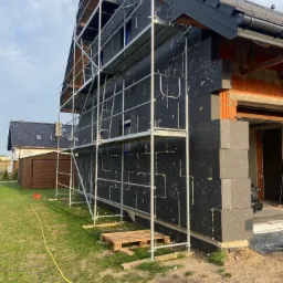 Dom w trakcie ocieplania elewacji styropianem grafitowym, widoczne rusztowanie, fragment niezabezpieczonej ściany z cegły oraz żółty wąż na trawie.