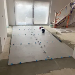 Układanie jasnych płytek podłogowych z użyciem niebieskich krzyżyków dystansowych w trakcie remontu, widoczne narzędzia glazurnicze i zabezpieczone folią schody.