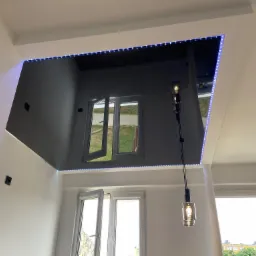 Nowoczesne wnętrze w Ozorkowie z podwieszanym, lustrzanym sufitem, obramowanym niebieskimi diodami LED, odbijającym okna i światło dzienne, uzupełnione wiszącą lampą z dekoracyjną żarówką.