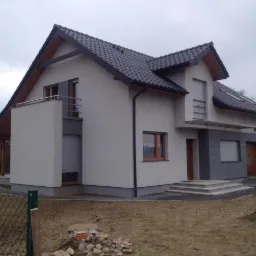 Nowoczesny dom jednorodzinny z szarą elewacją, ciemną dachówką i drewnianymi oknami, widoczny z perspektywy ogrodu w pochmurny dzień.