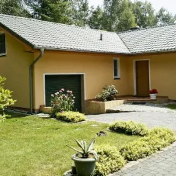 Parterowy dom z garażem i ogrodem. Żółta elewacja, szary dach, zielone drzwi garażowe. Brukowany podjazd, zadbany trawnik i rabaty kwiatowe.