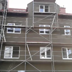 Budynek w trakcie tynkowania elewacji, widoczne rusztowania i świeżo nałożony tynk w kolorze szaro-zielonym, okna białe. Dach z czerwonej dachówki.