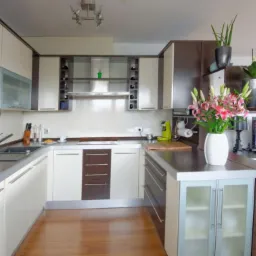 Nowoczesna kuchnia z białymi szafkami i drewnianymi akcentami, stalowym blatem, kwiatami w wazonie. Widoczne elementy wyposażenia i oświetlenie sufitowe.