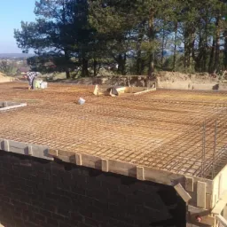 Budowa fundamentów zbrojonych siatką stalową i deskowaniem w Wejherowie, widoczny pracownik w tle i składowane drewno.