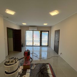 Wnętrze po malowaniu ścian na beżowo, z widocznymi narzędziami malarskimi, odkurzaczem i zabezpieczonymi folią oknami balkonowymi. Nowoczesne oświetlenie sufitowe.