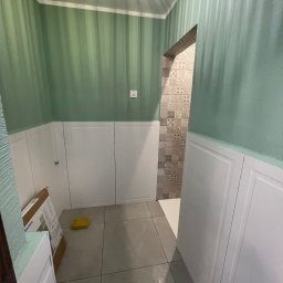 Wykończona łazienka z zielonymi ścianami w paski i białymi panelami. Widoczny fragment wejścia do kabiny prysznicowej z dekoracyjnymi płytkami. Na podłodze szare płytki.