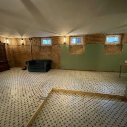 Pokój w trakcie remontu: odsłonięta cegła, fragment pomalowanej ściany, wzorzysta podłoga z ramą, antyczny kredens i sofa. Widoczne etapy prac wykończeniowych.