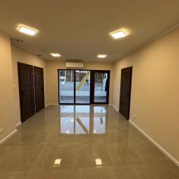 Patryk Gembara - Wykończone wnętrze: beżowe ściany, ciemne drzwi, błyszcząca, szara podłoga z płytek. Widoczne oświetlenie sufitowe i klimatyzacja. Przez szklane drzwi widok na zewnątrz.