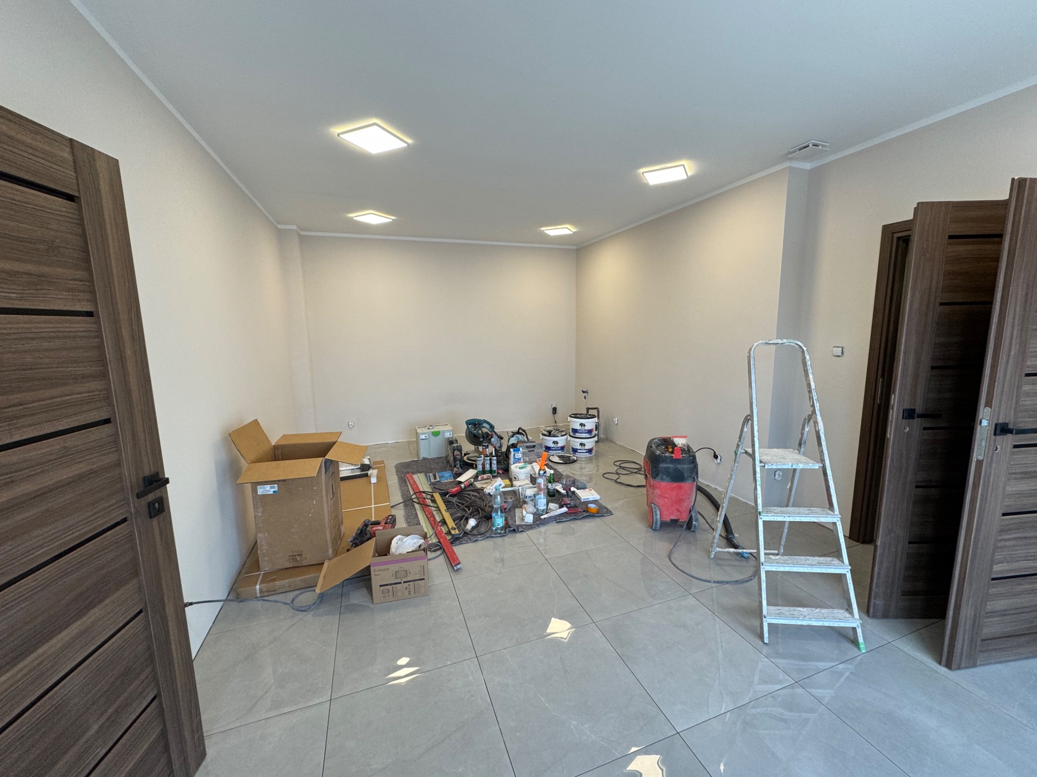 Remont pokoju w Ostravie: widok na przygotowane pomieszczenie z narzędziami, drabiną i materiałami malarskimi. Podłoga wyłożona szarą płytką, ściany przygotowane do malowania.