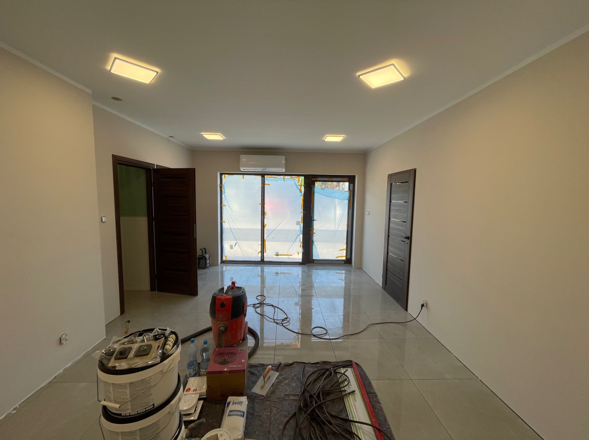 Wnętrze po malowaniu ścian na beżowo, z widocznymi narzędziami malarskimi, odkurzaczem i zabezpieczonymi folią oknami balkonowymi. Nowoczesne oświetlenie sufitowe.