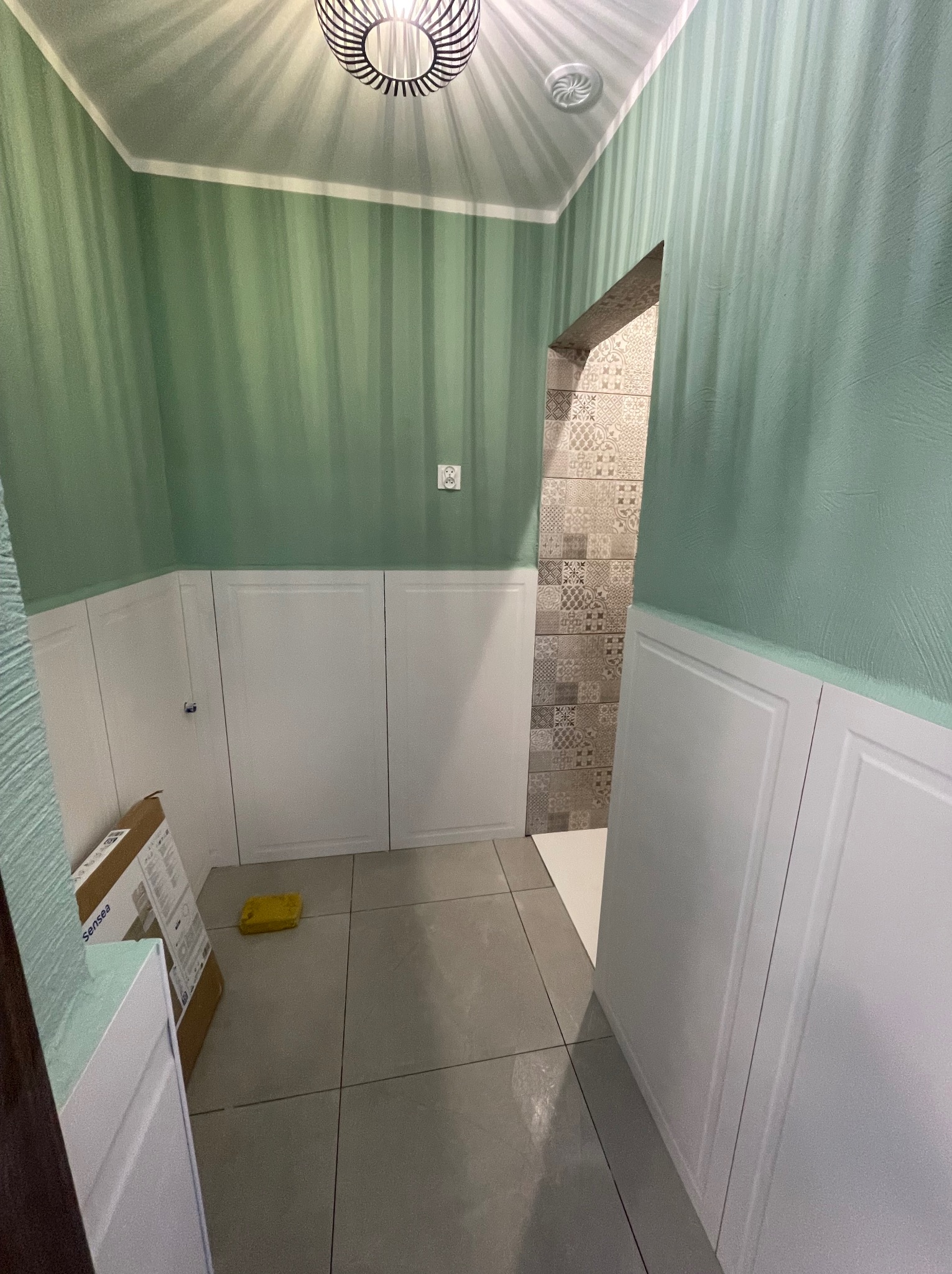 Wykończona łazienka z zielonymi ścianami w paski i białymi panelami. Widoczny fragment wejścia do kabiny prysznicowej z dekoracyjnymi płytkami. Na podłodze szare płytki.