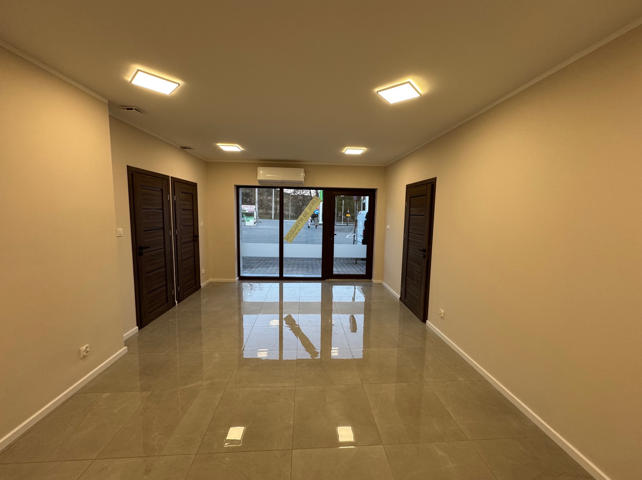 Wykończone wnętrze: beżowe ściany, ciemne drzwi, błyszcząca, szara podłoga z płytek. Widoczne oświetlenie sufitowe i klimatyzacja. Przez szklane drzwi widok na zewnątrz.