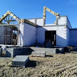 Budowa domu jednorodzinnego w stanie surowym otwartym: ściany z betonu komórkowego, drewniana konstrukcja dachu, widoczne bloczki budowlane i taczka.