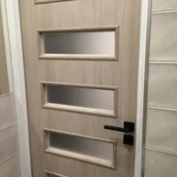 Jasne, drewnopodobne skrzydło drzwiowe z czterema prostokątnymi przeszkleniami z matowego szkła i czarną klamką, widoczne w kadrze fragmenty ościeżnicy i ściany z płytek.