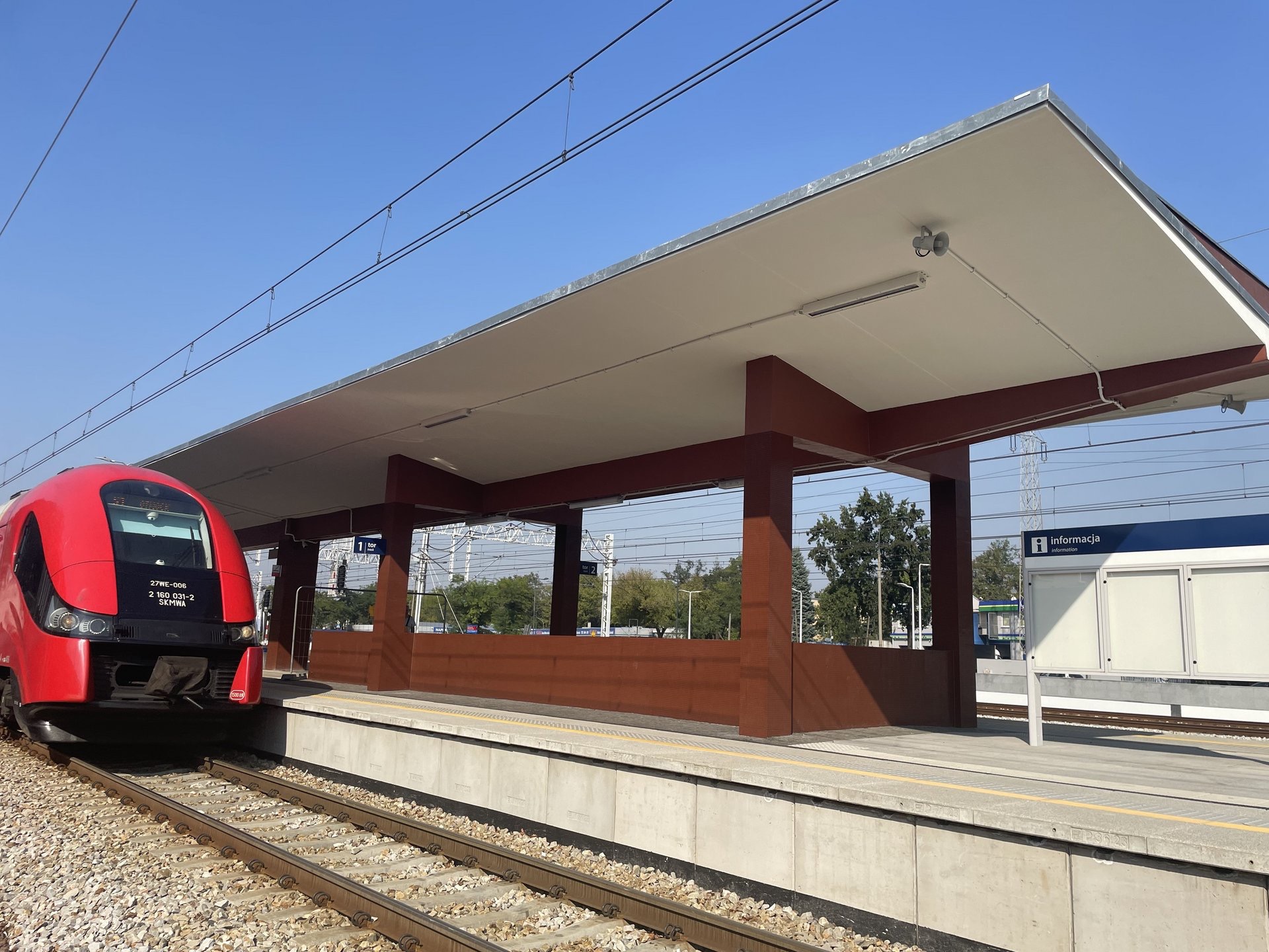 Nowoczesny peron kolejowy z czerwonym pociągiem, zadaszeniem wspartym na brązowych kolumnach i tablicą informacyjną na tle błękitnego nieba.