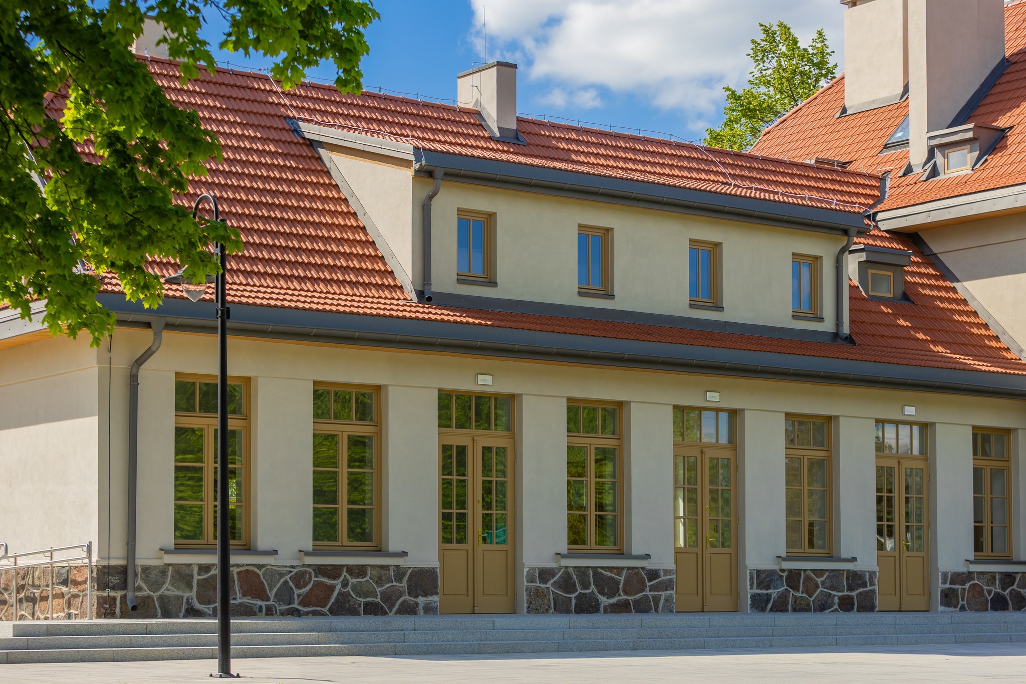 Odrestaurowany budynek z czerwoną dachówką i beżową elewacją, z kamiennym cokołem. Widoczne okna i drzwi w kolorze beżowym, oświetlenie uliczne i fragment drzewa.