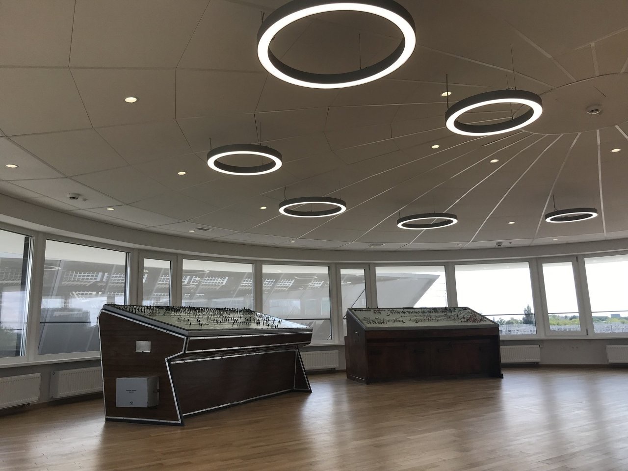 Jasne, okrągłe wnętrze z panoramicznymi oknami i dwoma dużymi modelami architektonicznymi na drewnianej podłodze, oświetlone nowoczesnymi lampami pierścieniowymi.