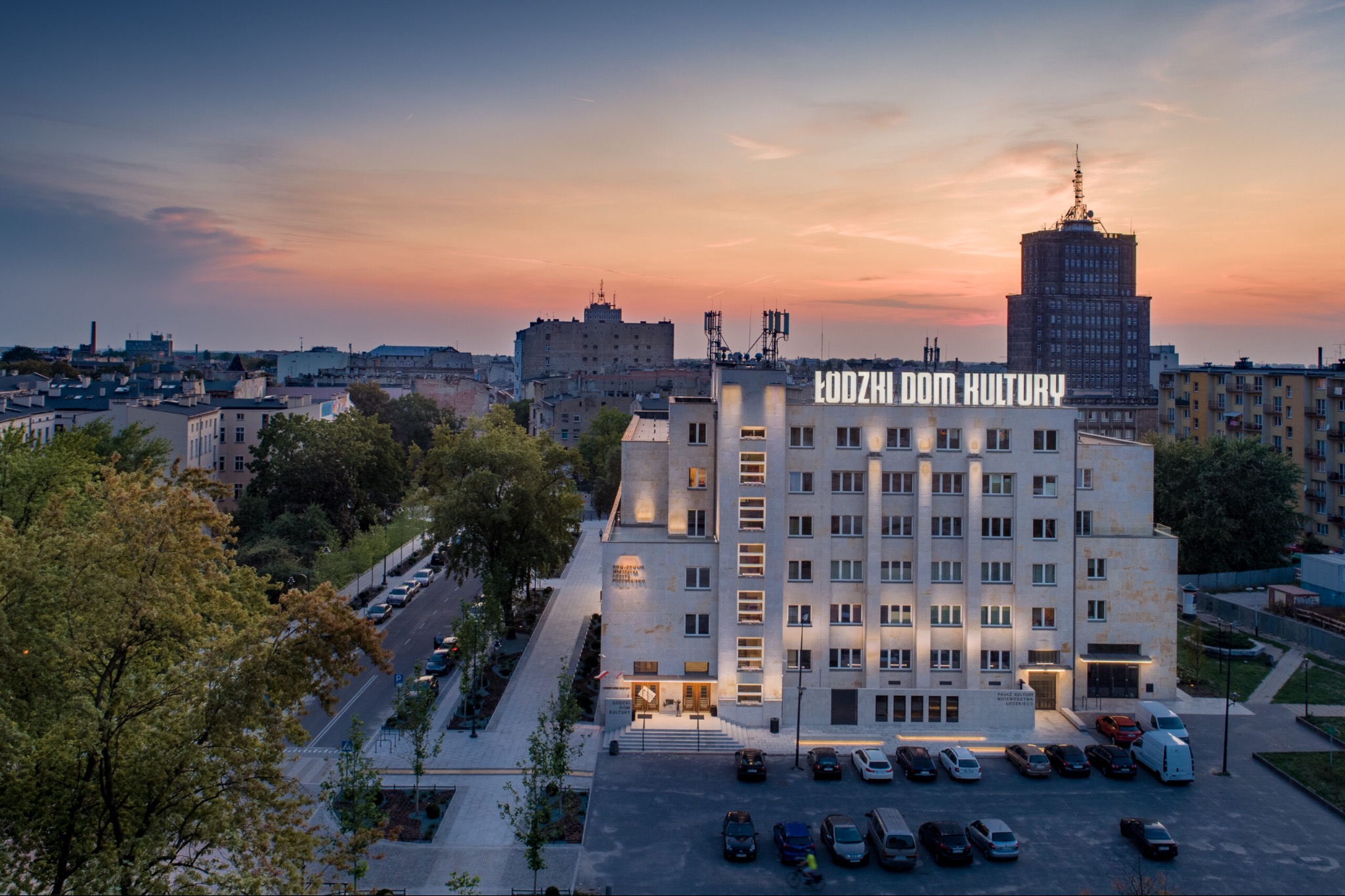 Odrestaurowany budynek Łódzkiego Domu Kultury oświetlony wieczornym światłem, z widoczną elewacją, parkingiem i panoramą miasta w tle.
